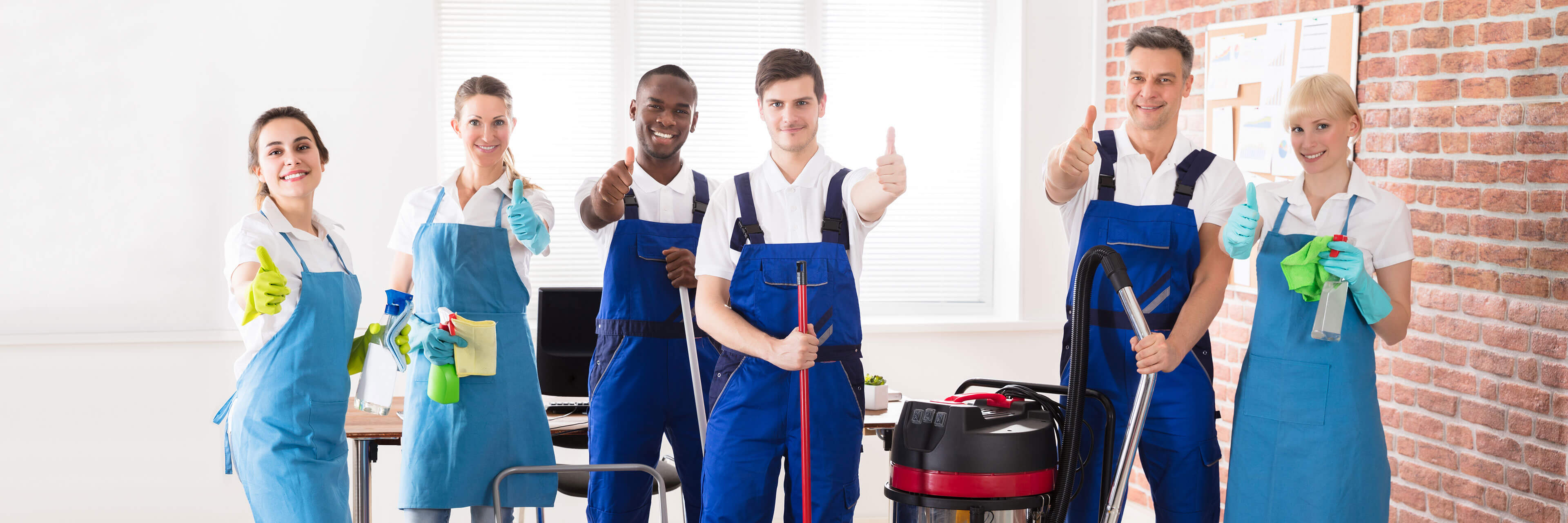 Ein Bild von sechs Reinigungskräften, die in Arbeitskleidung und mit Reinigungsmitteln in den Händen in einem Büro stehen. Dabei heben sie den Daumen nach oben.